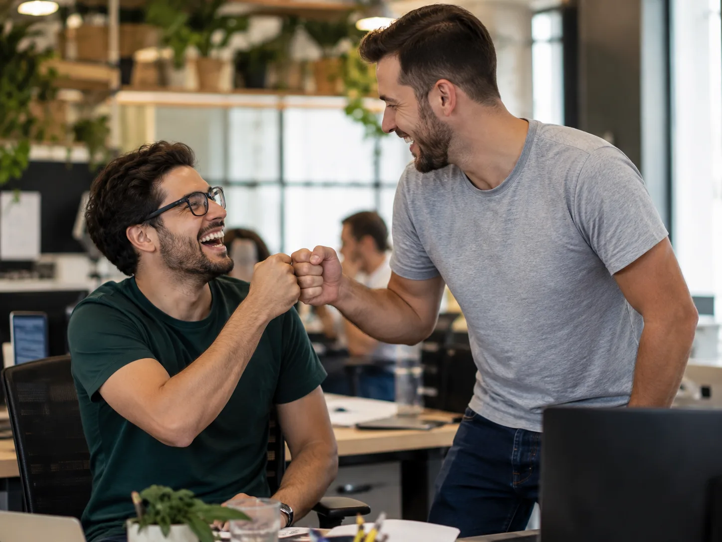 Dois colegas comemorando com um cumprimento de punhos em escritório aberto, sorrindo após atingir uma meta, com a equipe trabalhando ao fundo
